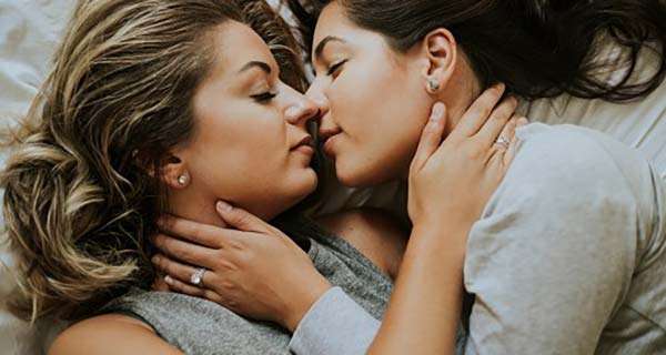 Dos lesbianas están acostadas juntas, mirándose de frente en una cama, abrazándose suavemente y a punto de besarse